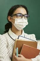 a young teacher wearing a face mask sits at a desk with books photo