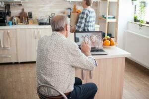 Happy senior man waving to his niece during a video conference with family using laptop in kitchen. Online call with daughter. Elderly person using modern communication online internet web techonolgy. photo