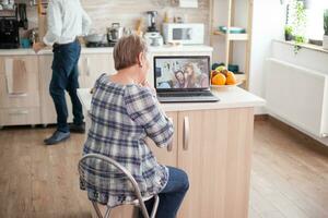 contento mayor mujer durante un vídeo conferencia con familia utilizando ordenador portátil en cocina. en línea llamada con hija y sobrina. mayor persona utilizando moderno comunicación en línea Internet web tecnología. foto