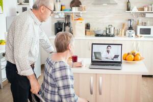 Doctor on telemedicine for senior couple. Video conference with doctor using laptop in kitchen. Online health consultation for elderly people drugs ilness advice on symptoms, physician telemedicine webcam. Medical care internet chat. photo