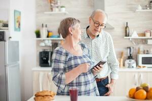 mayor mujer participación teléfono inteligente y compartiendo el receta con marido. caucásico mayor esposa y marido en cocina durante desayuno utilizando teléfono inteligente tecnología y Internet conexión ocio retirado mujer y hombre sonriente en domicilio, relajante. foto