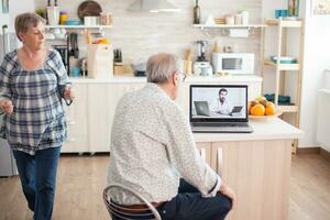 Senior couple during video conference with doctor using laptop in kitchen discussing about health problems. Online health consultation for elderly people drugs ilness advice on symptoms, physician telemedicine webcam. Medical care internet chat photo