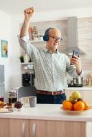 Happy mature adult listening music wearing headphones in kitchen during breakfast. Elderly retired person enjoying modern fun happy lifestyle, dancing relaxed, smiling and using modern technology photo