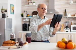 Happy senior man searching on tablet in kitchen during breakfast enjoying loisire time. Elderly retired person working from home, telecommuting using remote internet job online communication. photo