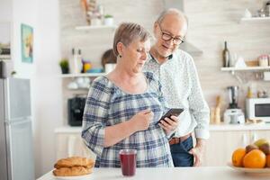 mayor Pareja utilizando moderno tecnología en su ocio tiempo. caucásico mayor esposa y marido en cocina durante desayuno utilizando teléfono inteligente tecnología y Internet conexión ocio retirado mujer y hombre sonriente en domicilio, relajante. foto