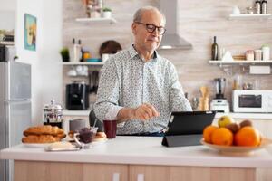 Elderly man smiling using tablet enjoying modern internet online technology, surfing on social media sitting in kitchen during breakfast. Authentic portrait of senior working from home at notepad photo