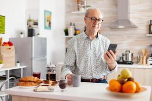 Happy old man surfing on social media using smartphone during breakfast sitting in kitchen smiling. Authentic portrait of retired senior enjoying modern internet online technology, searching, browsing photo