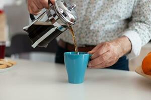 Man's hand pouring aromatic coffee into a blue cup from french press on a wooden table during breakfast. Elderly person in the morning drinking fresh brown espresso from vintage mug, filter relax refreshment photo