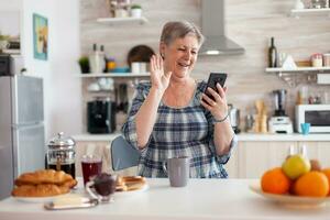 mayor mujer diciendo Hola mientras teniendo vídeo llamada con familia utilizando teléfono inteligente en cocina durante desayuno. mayor persona utilizando Internet en línea charla tecnología, tableta cámara web para virtual conferencia llamada foto