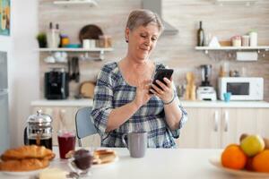 Relaxed elderly woman browsing on phone in kitchen during breakfast. Grandma using modern smartphone internet technology, online communication connected to the world, senior leisure time with gadget photo