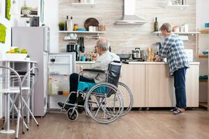 Handicapped man in wheelchair opening refrigerator and helping wife preparing breakfast in kitchen. Senior woman cooking for paralized husband, living with disabled man with walking disabilities photo