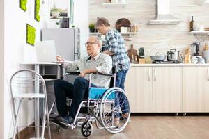 Paralysed elderly man in wheelchair typing on laptop working from home at computer in kitchen while wife is cooking breakfast. Handicapped businessman, entrepreneur paralysis for elderly retired male photo