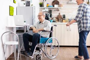Senior couple laughing using laptop, husband calling his wife near him during a video call with grandchildrens sitting in kitchen. paralyzedhandicapped old elderly man using communication techonolgy photo