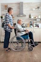 Wife pushing invalid senior dreamy man in wheelchair in kitchen, discussing and smiling, enjoying the beautiful morning together. Happy mature retired paralysed disabled positive male photo