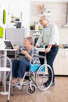 Paralysied senior wife in wheelchair working at tablet computer, showing to husband her project sitting in kitchen. Disabled handicapped old elderly person using modern communication online internet web techonolgy. photo