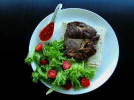 servicio blanco plato con crujiente frito carne, verduras, Delgado un pan y salsa en negro superficie foto