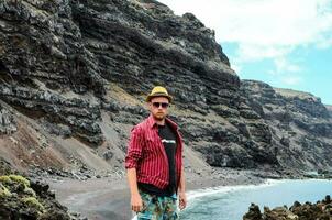 a man in a hat and sunglasses standing on a rocky beach photo