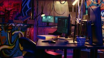 Dolly out shot of empty messy hackers base of operations with neon lights and graffiti drawings sprayed on walls. Ghetto hideout used by criminals to commit illegal activities using computers video