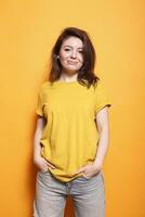 In studio, excited female person stands in front of isolated orange background. Joyful woman poses confidently, casually looking at the camera. Stylish adult with hands in pockets and a smile. photo