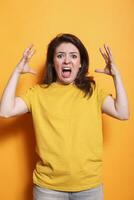 Caucasian woman with open mouth and raised arms, conveying anger and negativity. Annoyed lady in casual clothing stands in studio with orange background, screaming at the camera. photo