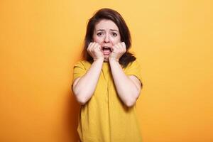 Terrified person with shocked expression standing over isolated orange background. While looking at camera, anxious woman is terrified and in distress with her hands on chin. photo