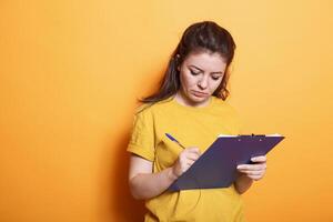 In the studio, young woman is using a pen to taking notes on paper. Focused individual signing project related paperwork on a notepad clipboard and writing in notebook files. photo