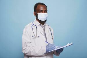 Portrait of black man wearing face mask and a lab coat while holding medical research documents. Male healthcare specialist posing for camera while grasping a pen and clipboard. photo