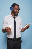 Busy black man with wireless headset browsing social media on mobile device while holding a beverage. Office worker on break listening to online music while drinking his cup of coffee. photo