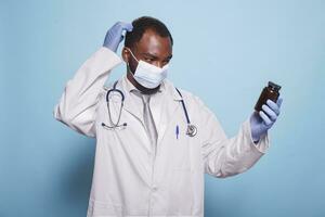 cuidado de la salud especialista vestido en médico protector engranaje mirando desconcertado a prescripción píldora botella. confuso negro médico vistiendo cara mascarilla, estetoscopio, y guantes agarre un analgésico envase. foto