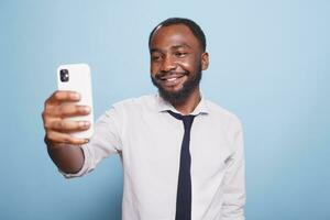 Businessman snapping a selfie with his smartphone front camera against isolated blue background. Happy independent freelancer video conferencing on his digital mobile device. photo