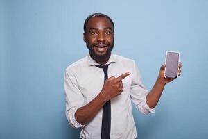 Enthusiastic black man pointing at smartphone blank screen showing mobile feature of mobile game or app. Excited individual using index finger to enhance features of digital device. photo