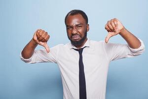 Annoyed black man showing thumbs down gesture on camera, making disagreement and disapproval sign in studio. Displeased individual posing over blue background with negative symbol. photo