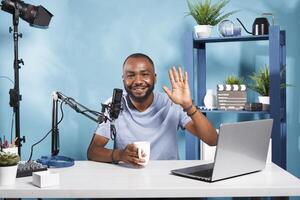 Smiling blogger speaking and greeting subscribers while streaming online on channel. Cheerful african american vlogger drinking coffee while waving hi and creating web content portrait photo