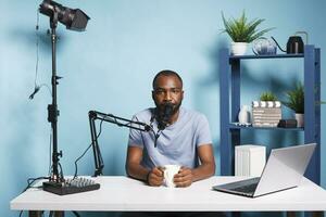 African american blogger recording video for internet channel while drinking coffee and looking at camera. Man vlogger live streaming for online podcast and speaking in microphone photo