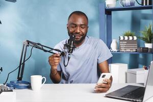 Smiling vlogger answering questions while holding smartphone and streaming online. Web content creator reading news on smartphone while recording video for internet channel photo