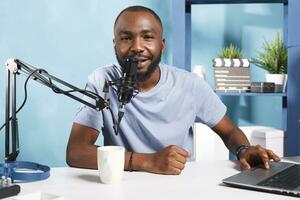 Smiling vlogger speaking in microphone, using laptop and looking at camera while recording video for internet channel. Influencer blogger talking in mic while streaming online portrait photo