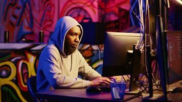 Portrait of african american hacker in abandoned warehouse using phishing technique that tricks users into revealing sensitive information. Hooded cybercriminal stealing passwords video