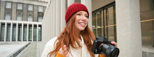 sonriente pelirrojo niña fotógrafo, tomando imágenes en ciudad, hace fotos al aire libre en profesional cámara