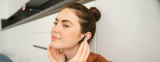 Close up portrait of smiling cute young woman, listens to music in wireless headphones, enjoying quality of sound in new black earphones, sitting on floor at home photo