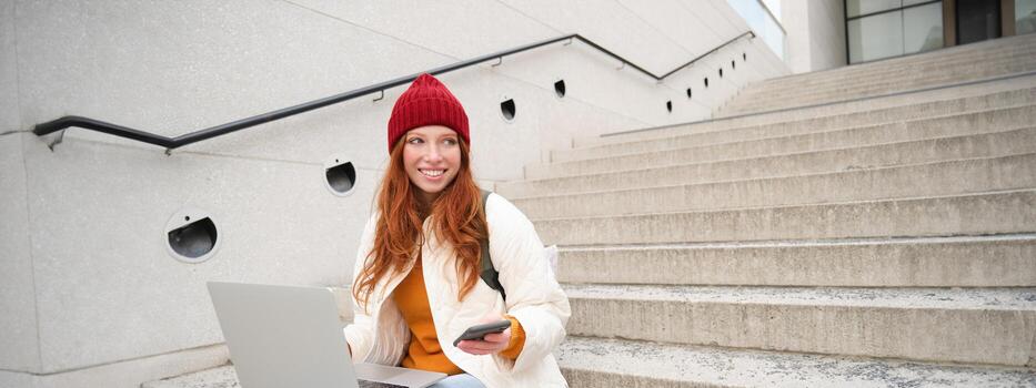 Smiling redhead woman with mobile phone and laptop, sitting on stairs outside building, connects to public wifi, using smartphone and computer photo