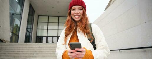 Happy girl student in red hat, holds smartphone, tourist looks at map app on her phone, explores sightseeing, texts message, looks for couchsurfing, rents place to stay online photo