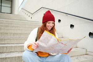 joven sonriente pelirrojo chica, turista se sienta en escalera al aire libre con ciudad papel mapa, mirando para dirección, viajero mochilero explora ciudad y mira para Turismo foto