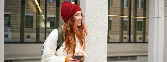 Stylish urban girl using mobile phone app, standing in city, waiting for taxi, looking at smartphone application, texting message photo