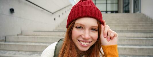 Stylish redhead girl in warm red hat, smiling relaxed, sitting with backpack on stairs near building, waits for someone outdoors photo