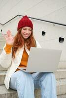 retrato de contento joven mujer, pelirrojo niña con computadora portátil, olas mano y dice Hola en vídeo charlar, tiene conversacion en computadora solicitud, negociaciones a alguien foto