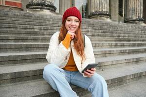 Portrait of young urban girl in red hat, sits on stairs near museum, holds mobile phone, connects to public wifi and surfs net, uses smartphone apps photo