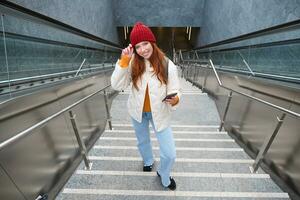 Portrait redhead girl tourist, goes up stairs with smartphone, follows route on mobile phone app, holds backpack and smiles photo