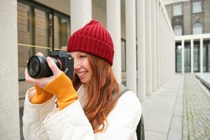 sonriente pelirrojo niña fotógrafo, tomando imágenes en ciudad, hace fotos al aire libre en profesional cámara