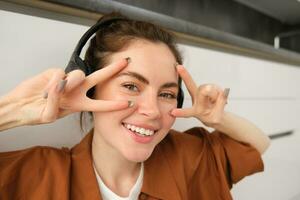 cerca arriba de despreocupado joven mujer, riendo y sonriente, demostración paz, vsign gesto, escuchando a música en inalámbrico auriculares foto