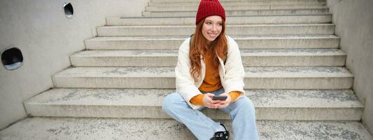 Hipster ginger girl, redhead woman sits on stairs with smartphone, waits for someone and messages on social media on mobile phone app photo